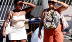 Duas mulheres cobrem os rostos com as mãos, tentando se proteger do sol, ambas vestem blusas de alcinhas e carregam bolsas e mochila