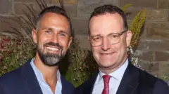 Jens Spahn (jobbra) szemüvegben, sötét zakóban, fehér ingben és piros nyakkendőben áll férje, Daniel Funke mellett, aki szakállt visel, valamint sötétkék zakót és kék inget.
