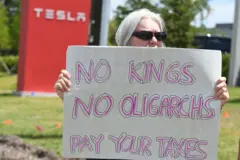 Uma mulher segura um cartaz em frente a uma concessionária da Tesla com os dizeres: “Sem reis, sem oligarcas — paguem seus impostos"