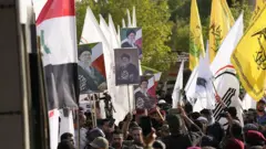 İran ve Irak bayrakları taşıyan kişiler, 29 Temmuz 2026'da Irak'ın Bağdat kentindeki çeşitli noktalarda ABD ve Suudi güçleri tarafından gerçekleştirildiği bildirilen hava saldırılarında hayatını kaybeden Haşdi Şabi (PMF) mensupları için düzenlenen cenaze törenine katıldı. Kalabalığın ellerinde PMF bayrakları ve Hamaney dövizleri görülüyor. 