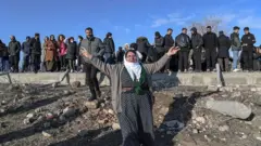 Beyaz baş örtülü bir kadın kollarını açmış. Arkadada çoğu erkek kalabalık sınır tellerinin yanında duruyor. Gökyüzü mavi