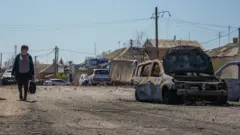 Uma mulher caminha por uma rua ensolarada, carregando uma sacola de compras, ao lado de um carro destruído por uma bomba.