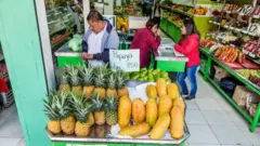 Interior de mercado, com frutas e legumes à venda, funcionários e clientes