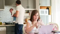 Bir baba çocuğu tutarken, anne mutfakta bilgisayarın başında.