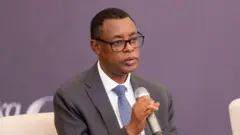 James Kabarebe yicaye arimo kuvugira mu ndangururamajwi ayifashe mu kiganza. Yambaye ikositimu yijimye, ishati y'umweru na karuvati irimo amabara mato y'ubururu n'umweru. 