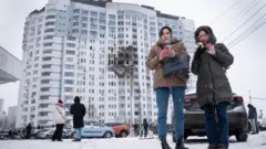 厚着の冬服に身を包んだ2人が手前に立っている。地面には雪が積もっている。背後の高層住宅は、中央に爆発物の直撃を受けた大きな黒い焼け跡がある