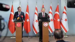 Yılmaz ve Üstel kürsüden konuşuyor. Arkalarında Türk ve Kuzey Kıbrıs bayrakları var.