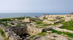 Руїни античного міста Херсонес біля морського узбережжя. Серед кам’яних фундаментів і залишків стародавніх споруд видно доріжки та кількох відвідувачів, а на задньому плані простягається море під ясним небом.
