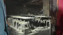 Mesas e cadeiras estão cobertas de cinzas dentro do local, que apresenta grandes danos causados pelo fogo. A foto foi tirada através de uma moldura de porta