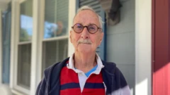 Jeff Stein, de óculos e bigode branco, veste uma camisa polo listrada e olha para a câmera em frente a uma casa