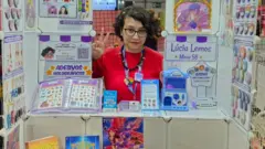 A ilustradora Lúcia Lemos, mulher jovemaviãozinho pixbetpele clara posa atrásaviãozinho pixbetestande. na frente, livros e desenhosaviãozinho pixbetpersonagens  