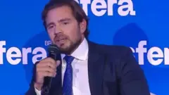 Vorcaro fala ao microfone de terno e grava azul, em frente a um painel azul