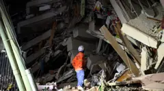 A rescue worker in a bright orange top stares at a collapsed building, with a vast mound of rubble, blocks of concrete and twisted steel in view.