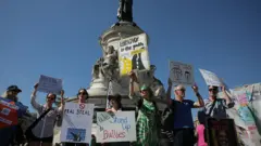 Uma fila de pessoas segura cartazes que dizem "nós enfrentamos os que fazem bullying", "parem o verdadeiro roubo do Doge" e "a empatia é o caminho" em uma rua de Paris