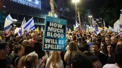 Pessoas na rua durante manifestação, com bandeiras de Israel e um cartas perguntando, em inglês, "quanto sangue ainda será derramado?".