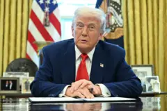 Trump arkasında Amerikan bayrağı ve başkanlık arması ile masada oturmuş, elinde bir kalem ile önündeki kağıt üzerinde ellerini kavuşturmuş
