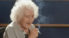 A francesa Jeanne Calment no seu 117º aniversário. Ela tem cabelos brancos ondulados e segura um cigarro aceso na mão, enquanto olha para longe da câmera, à direita. Ela veste um cardigã cinza e uma camisa preta e branca.