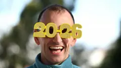 「2026」のかたちをしたフレームのめがねをかける男性が、笑顔でカメラを見ている