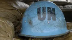 A UN blue helmet rests on sandbags next to a peacekeeper on duty.