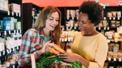 Duas mulheres conversam e escolhem cenouras no supermercado