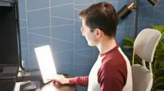 Jovem de óculos, sentado, olha para uma lâmpada usada para tratamento da depressão sazonal