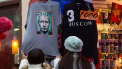 Duas mulheres usando chapéus examinam uma camiseta cinza com a capa da revista Time estampada, que está pendurada na porta de uma loja de souvenirs.