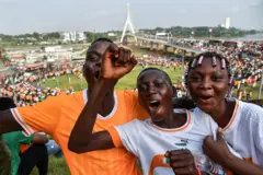 De jeunes enfants t-shirt orange et deux en blanc aux couleurs de la Côte d'Ivoire, en train de crier devant la caméra, une foule sur un pont et dans un espace vert est visible en arrière-plan