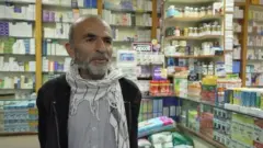 یکی ه از بیماران دارای  التهاب روده  در داخل یک دواخانه دیده می‌شود او ریش سفید دارد و در پشت سرش قفصه‌های دوا است 
