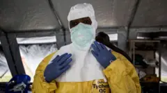 Man wear yellow overal to protect imsef, e wear blue gloves and use white nosemask while e dey stand for makeshift tent