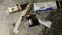 A imagem mostra diferentes itens associados ao uso de esteroides anabolizantes e testosterona dispostos sobre uma superfície de concreto. Entre eles, há duas caixas de medicamentos — uma identificada como Primobol-100 e outra como Testoviron Depot 100 mg/ml —, um frasco de vidro com tampa vermelha e duas seringas embaladas individualmente em plástico estéril. A composição destaca os produtos e os materiais utilizados para aplicação injetável dos hormônios.