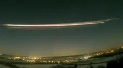 A photograph using 30-second exposure showing the Falcon 9 upper stage re-entering the atmosphere above Berlin, Germany. A long streak of bright yellow and white is seen in the dark night sky, above a landscape of dark field and bright lights from the city.