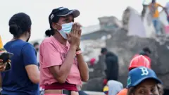 Žena u roze majici sa maskom na licu stoji skupljenih ruku