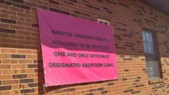 Uma placa rosa pendurada em um prédio de tijolos diz: “A Bristol Women’s Health tem a honra de ser a única clínica de aborto oficialmente designada de Bristol”.
