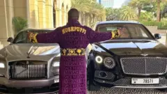 Hushpuppi devant des voitures de luxe.