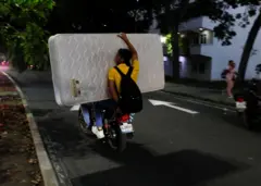 Dos personas viajan en una moto cargando un colchón entre ambas.