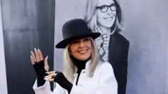 Dajen Kiton ima crni šešir na glavi smeje se i nosi beli kaput. Pokazuje ruke na kojima ima crne rukavice bez prstiju