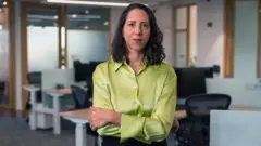 Laura Carvalho veste uma blusa verde de cetim e mangas longas, e tem os braços cruzados. Ela olha para a câmera com um sorriso discreto