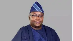 Minisita fun ọrọ agbara ni Naijiria; Adebayo Adelabu. O wọ agbada alawọ aro, de fila awọ kan naa, o si fi jigi soju bo ti n rẹrin-in musẹ ninu aworan yii.