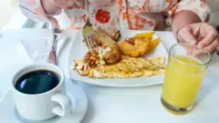 Persona desayunando con café, jugo y un plato con omelette, pan y frituras.