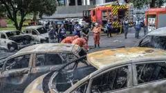 Bomberos y policías trabajan junto a vehículos quemados en el lugar del accidente de helicóptero en el barrio Recreio dos Bandeirantes en Río de Janeiro, Brasil, el 14 de junio de 2026.