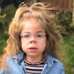Sophia Scott, una niña pequeña, está mirando a la cámara. Tiene el cabello largo y rubio, con una parte recogido en la parte superior con una goma. Lleva gafas de montura rosa pálido y una camisa o chaqueta vaquera, con una camiseta de rayas rosas, grises, negras y blancas debajo.