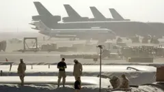 Önde askeri personal yükseltiler üzerinde geride uçaklar sislerin ardından görünüyor. 