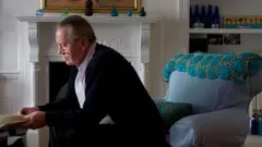 Chuck Feeney sentado de perfil em sala de estar, olhando para livro