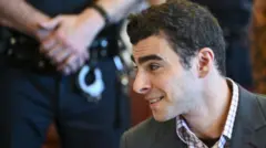 Улыбающийся молодой мужчина сидит за столом, на заднем плане не в фокусе виден силуэт человека в форме, держащего наручники