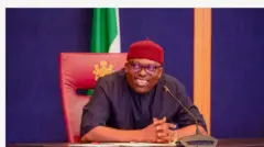 Aworan Siminalayi Fubara, Gomina ipinlẹ Rivers to ṣẹṣẹ pada sipo. O de fila pupa, o wọ ẹwu alawọ dudu, o n rẹrin-in bo ti n wo ẹgbẹ, o ki ika rẹ bọnu ara wọn lorii tabili to kawọ naa si, ẹrọ gbohun-gbohun kan wa niwaju rẹ