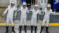 Cuatro astronautas con trajes espaciales blancos se toman de las manos y las alzan en señal de celebración a la entrada del Centro Espacial Kennedy.