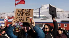 抗議デモに多くの人が集まっている。プラカードやグリーンランドの旗を掲げている人もいる。背景には雪が積もった山が見える