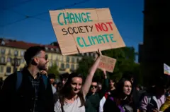 Vários jovens em uma manifestação. Um dos participantes segura um cartaz com os dizeres: Mude a sociedade, não o clima.