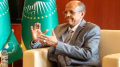 Mahmoud Ali Youssouf arongoye Inama nyobozi y'Ubumwe bwa Afrika ariko aravuga, yambaye amarori n'ikositime isa n'umunyota, ishati igomba kwera n'ikaruvati ifise utudomagudomagu, imbere yiwe ku ruhande hari amabendera y'Ubumwe bwa Afrika.