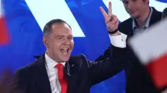 Karol Nawrocki stands on stage, gesturing the peace sign with his hand, wearing a dark suit, white shirt and red tie. A Polish flag is out of focus in the foreground.
